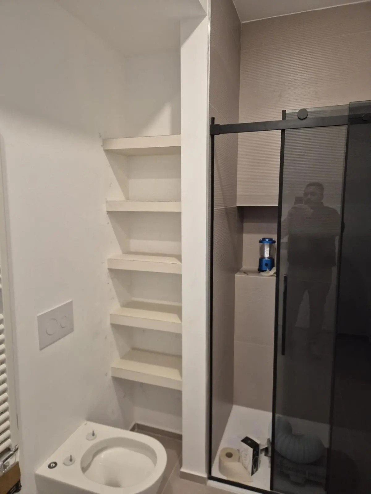Nicchia in cartongesso con mensole bianche accanto a doccia in vetro nero e vaso sanitario.