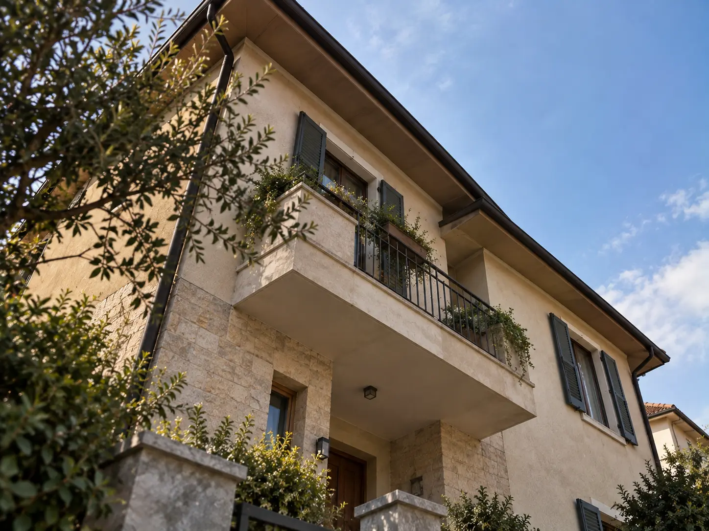Facciata color crema di una casa a due piani, con balcone fiorito, persiane scure e rivestimento in pietra.