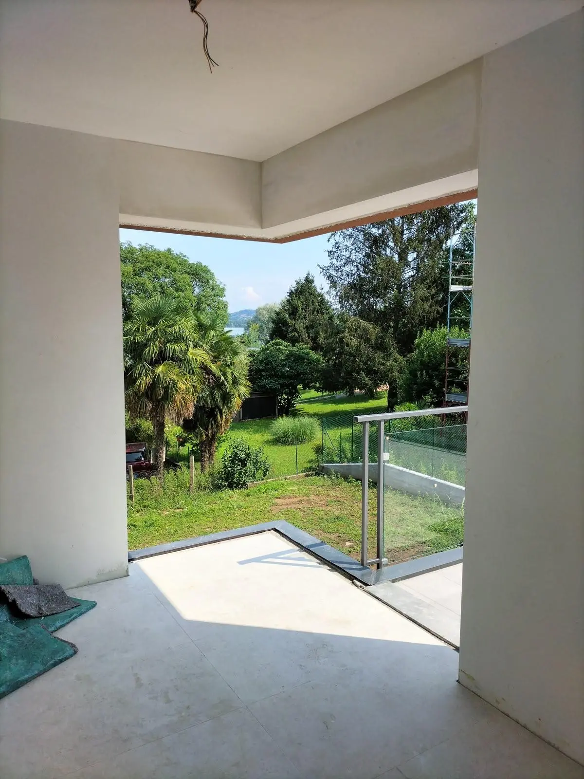 Interno in ristrutturazione aperto su un balcone con parapetto in vetro e vista sul giardino alberato.