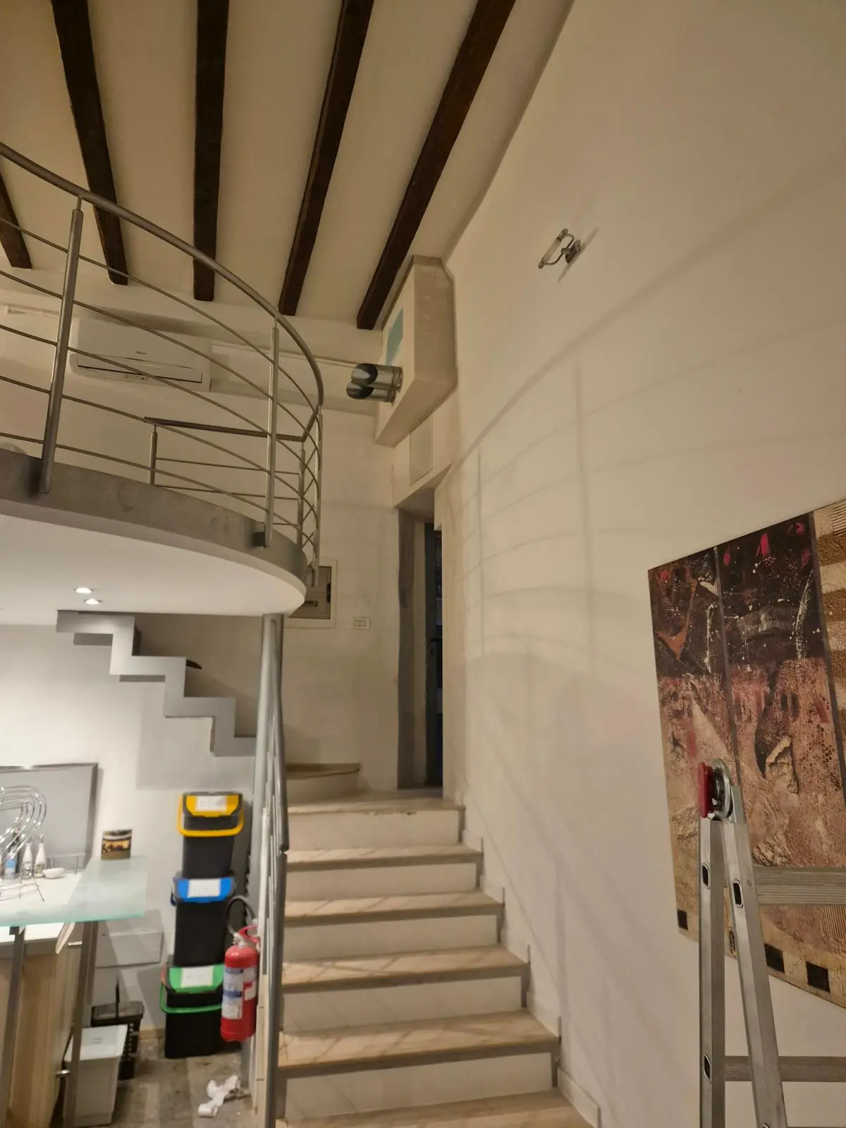 Scala interna in pietra con ringhiera metallica curva, soppalco, travi a vista e scala portatile.