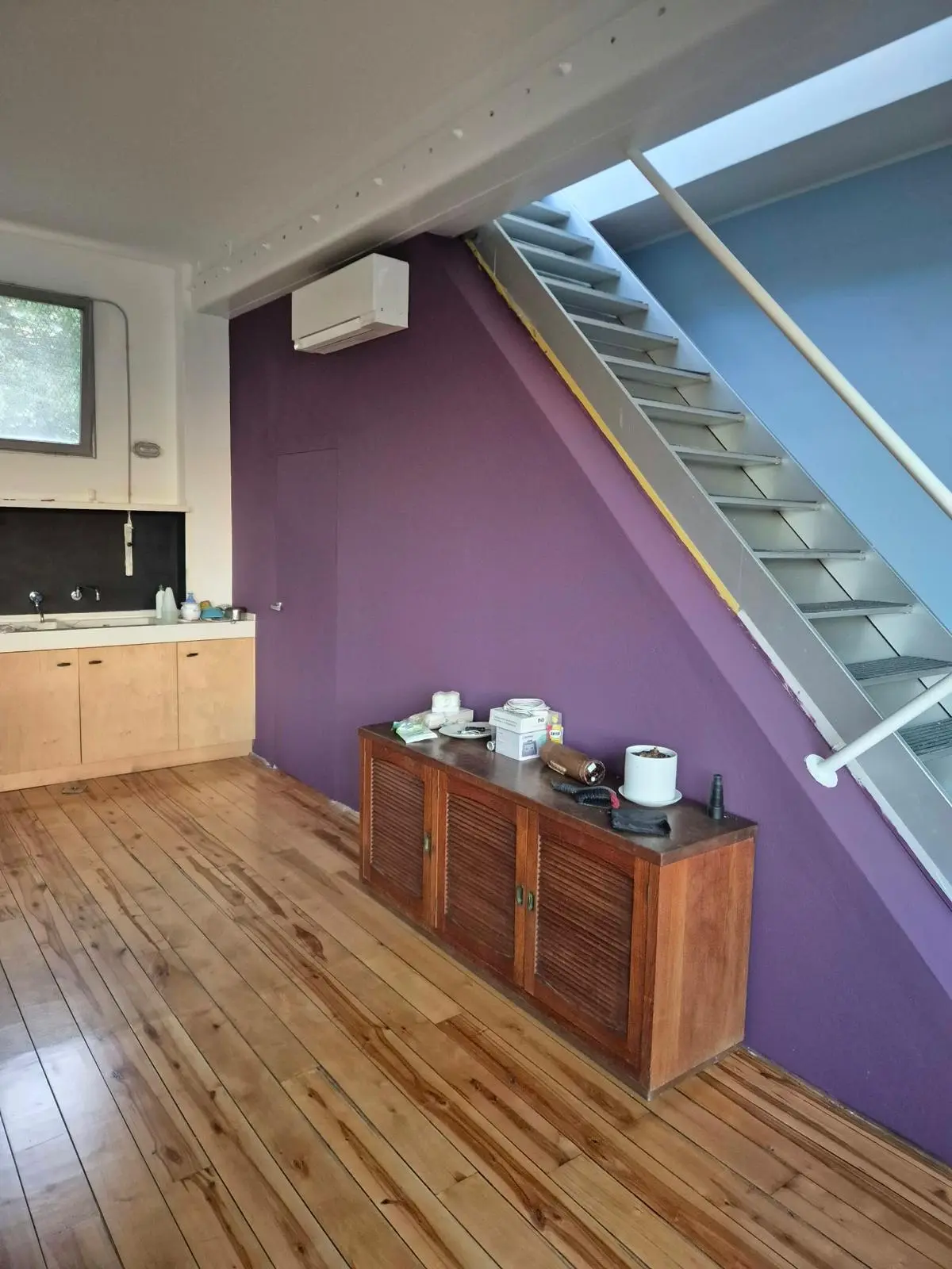Scala grigia accanto a una parete viola, con credenza in legno, pavimento a listoni e piccolo angolo cucina.
