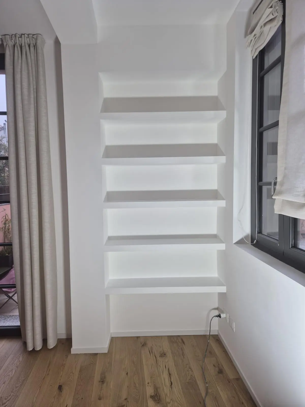 Libreria a parete bianca con cinque ripiani incassati, tra due finestre con tende e pavimento in legno.