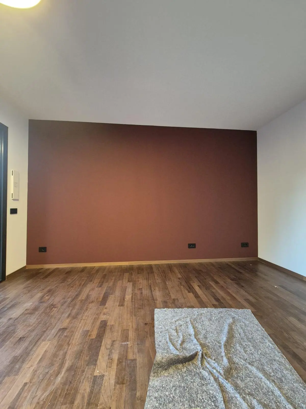 Stanza vuota con parete color terracotta, parquet in legno e tappeto grigio ripiegato sul pavimento.