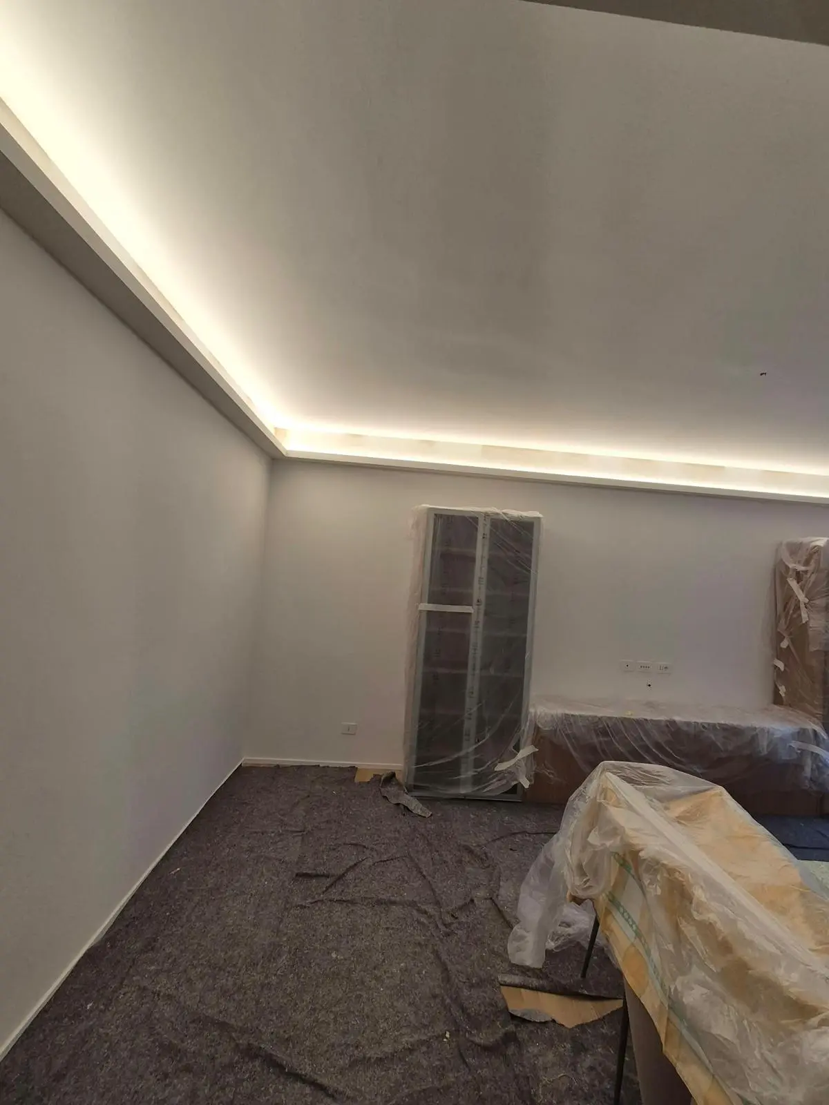 Stanza in ristrutturazione con illuminazione perimetrale a soffitto e mobili coperti da teli protettivi.