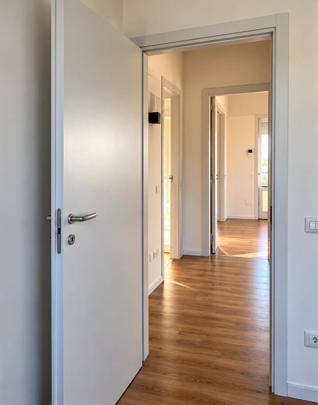 Porta interna bianca aperta su un corridoio luminoso con pavimento in legno e infissi coordinati.